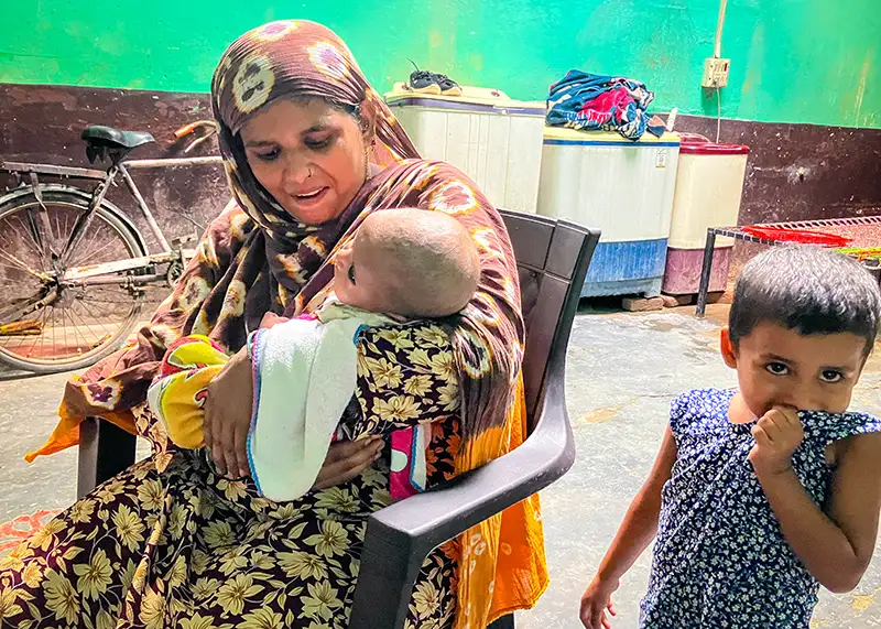 A seated woman cradles and infant, a toddler standing by her side.