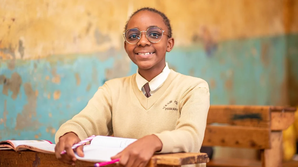 A teenaged girl wearing a school uniform and wire-rimmed glasses sits at a desk.
