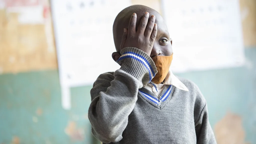 A boy in a school uniform covers his right eye with his hand. A Snellen eye chart is visible in the background.