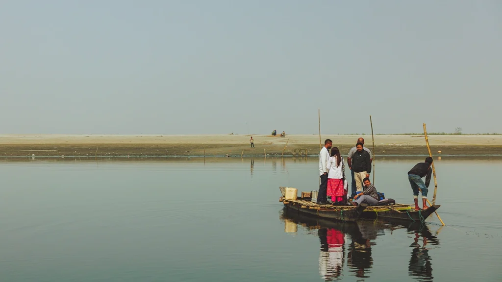 Six people sit or stand on a small boat that is crossing a wide river.