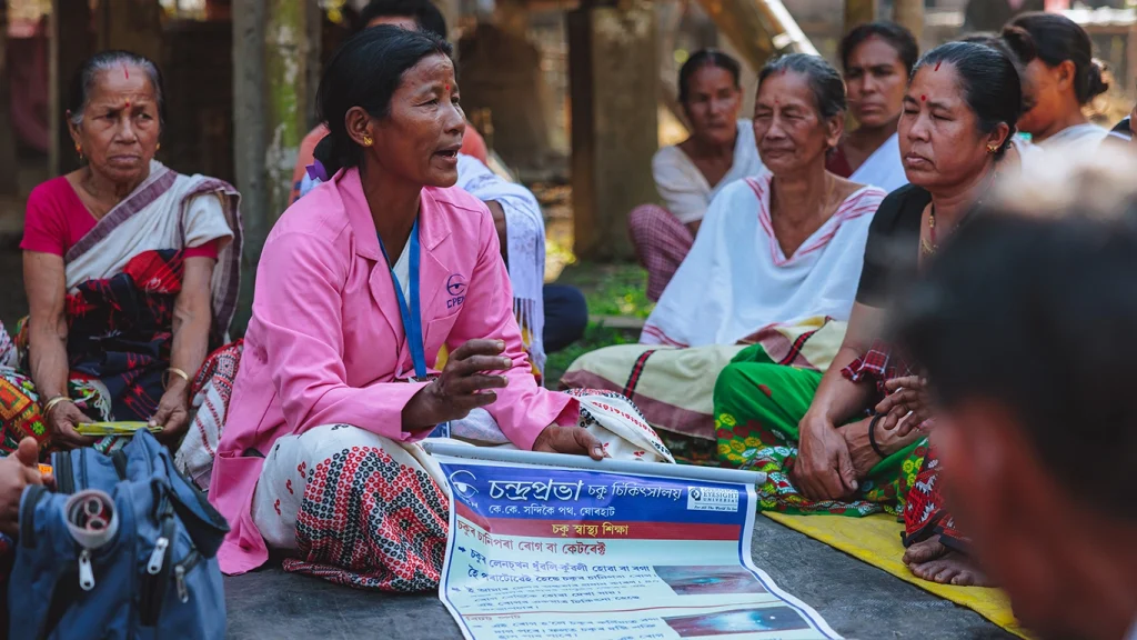 A woman in a pink medical coat shows a poster about eye health to a group of men and women gathered around her.
