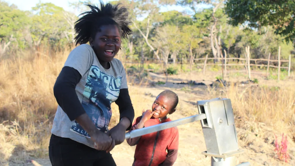 A teenage girl pushes down on the handle of a hand pump, while a child in the background smiles at the camera.