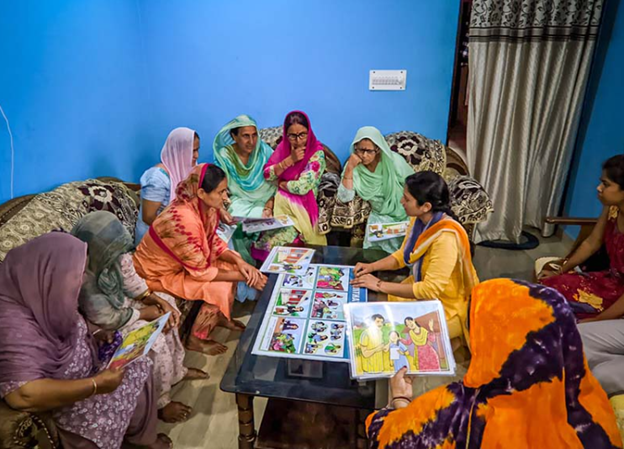 A group of women sit at a coffee table, discussing a number of drawings spread out before them.