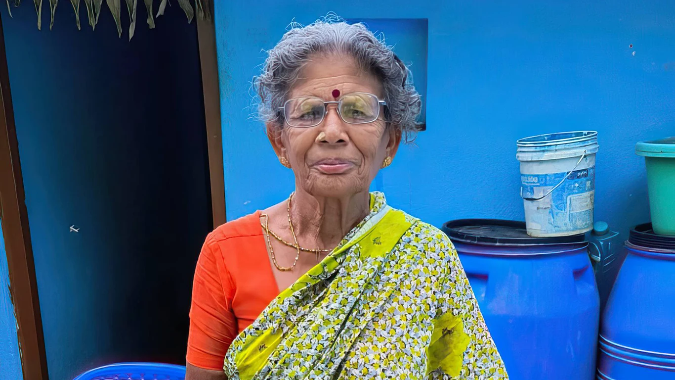 A woman wearing a yellow sari with orange blouse poses with her new eyeglasses on in front of a bright blue wall.